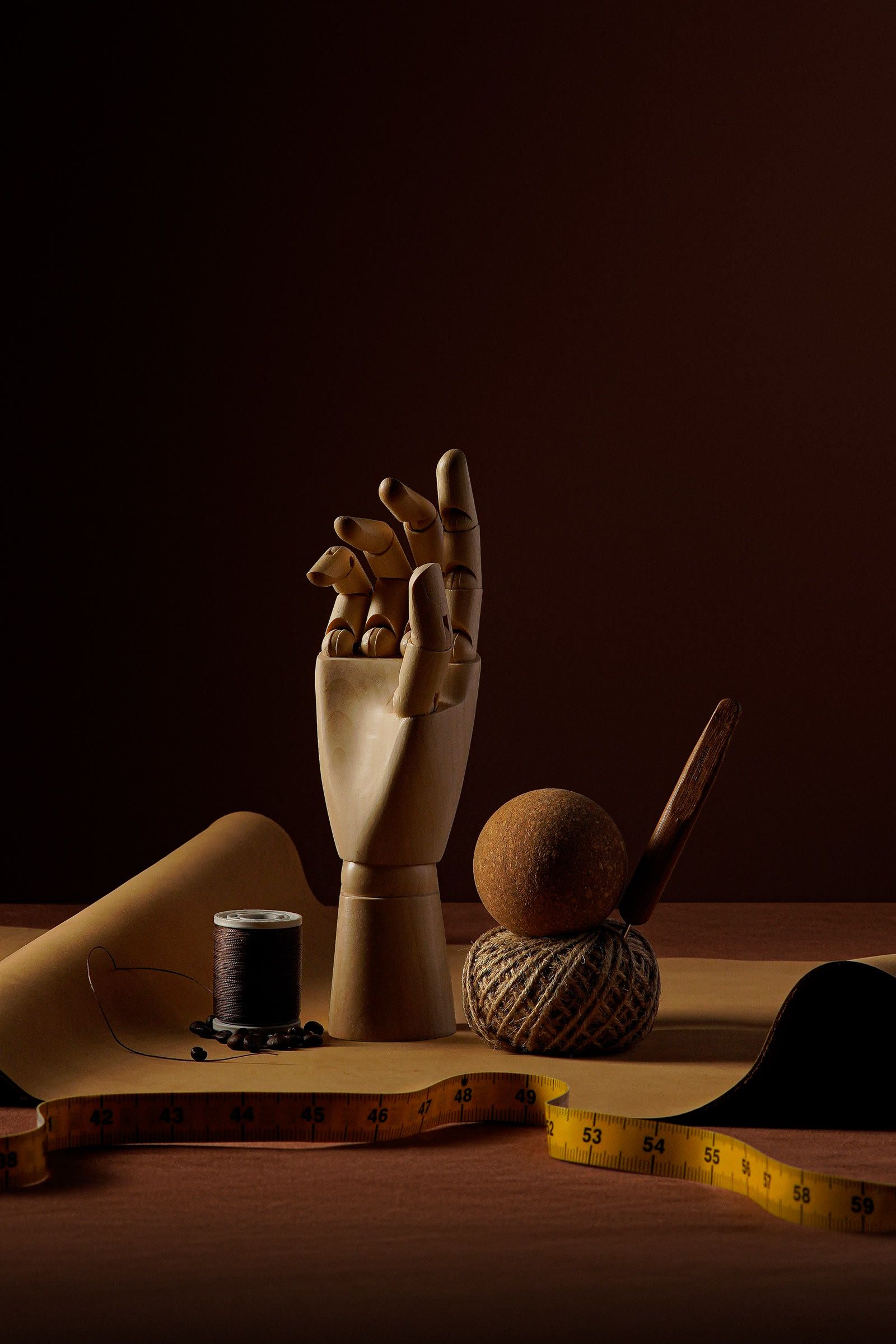 A wooden hand, a spool of thread, a ball of twine and a yellow tape measure on kraft paper.